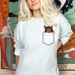 May include: A light gray sweatshirt featuring a graphic of a brown rat peeking out of a pocket. The pocket is outlined in black. The rat has pink ears and a detailed face. The sweatshirt is a casual style.