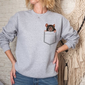 May include: A light gray sweatshirt featuring a black pocket design with a realistic illustration of a brown rat peeking out. The rat has dark eyes and a detailed fur pattern. The sweatshirt has long sleeves and a crew neck.