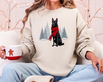 Op de afbeelding: Crèmekleurige sweatshirt met een afbeelding van een zwarte hond met een rode sjaal, tegen een achtergrond van gestileerde dennenbomen. De sweatshirt heeft een relaxte pasvorm. Een rood-witte mok met een peperkoekmannetje wordt in de hand gehouden.
