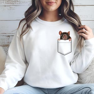 May include: White sweatshirt featuring a graphic of a brown rat peeking out of a pocket. The rat has dark brown fur and pink ears and nose. The pocket is outlined in black. The sweatshirt is a solid white color.
