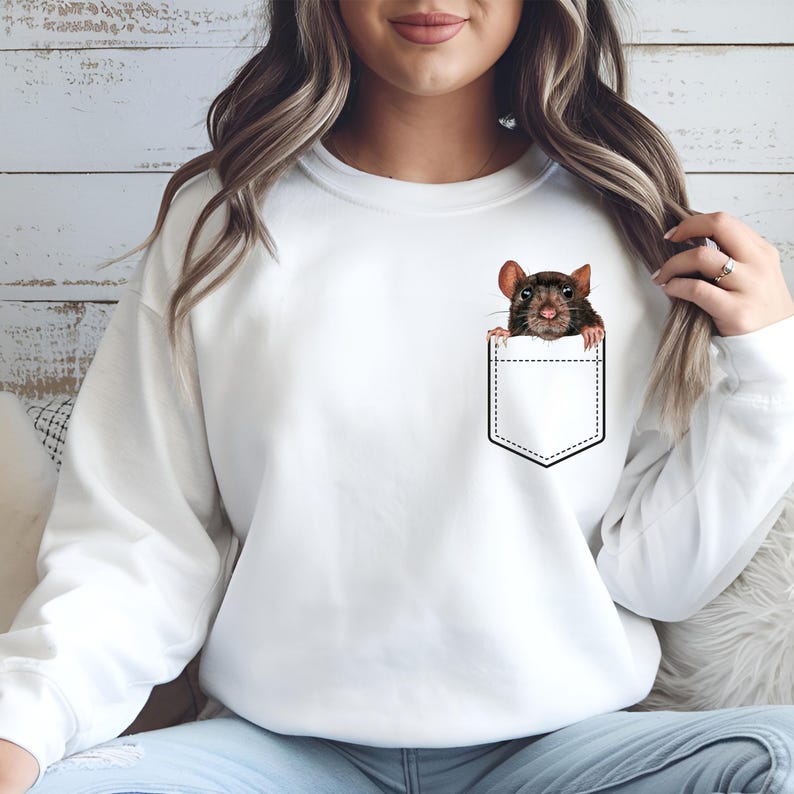 May include: White sweatshirt featuring a graphic of a brown rat peeking out of a pocket. The rat has dark brown fur and pink ears and nose. The pocket is outlined in black. The sweatshirt is a solid white color.