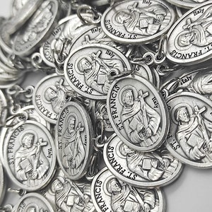 Può includere: Un mucchio di ciondoli ovali d'argento con l'immagine di San Francesco d'Assisi. Ogni ciondolo ha le parole "St. Francis" e "Italia" stampate su di esso.