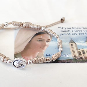 Puede incluir: Un brazalete anudado beige con cuentas plateadas y un dije plateado con una imagen de la Virgen María. El brazalete se muestra en una tarjeta con una cita de la Virgen María: "Si supieras cuánto te amo, llorarías de alegría." y una imagen de una iglesia.