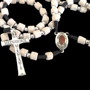 May include: White wooden bead rosary with a silver cross and a silver oval charm with a red stone. The cross has the words "Medjugorje" and "Mir" inscribed on it.