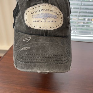 May include: Distressed grey baseball cap with a mesh back. The front has a patch with the text "Rattlesnake Island" and coordinates 43.56° N 71.28° W. The cap has a pre-curved peak and distressed details.