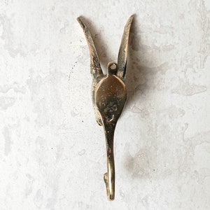 May include: A gold-toned metal hook shaped like a bird with its wings spread out. The hook is mounted on a white wall.