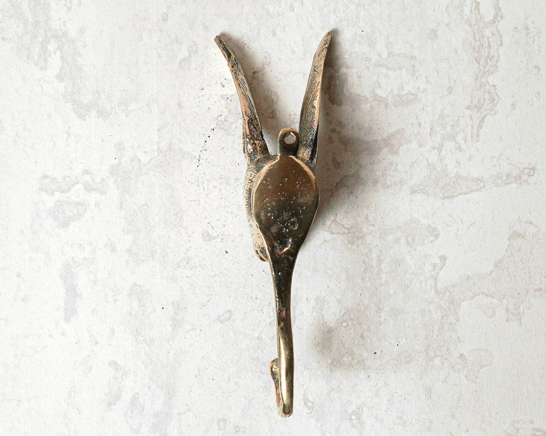 May include: A gold-toned metal hook shaped like a bird with its wings spread out. The hook is mounted on a white wall.