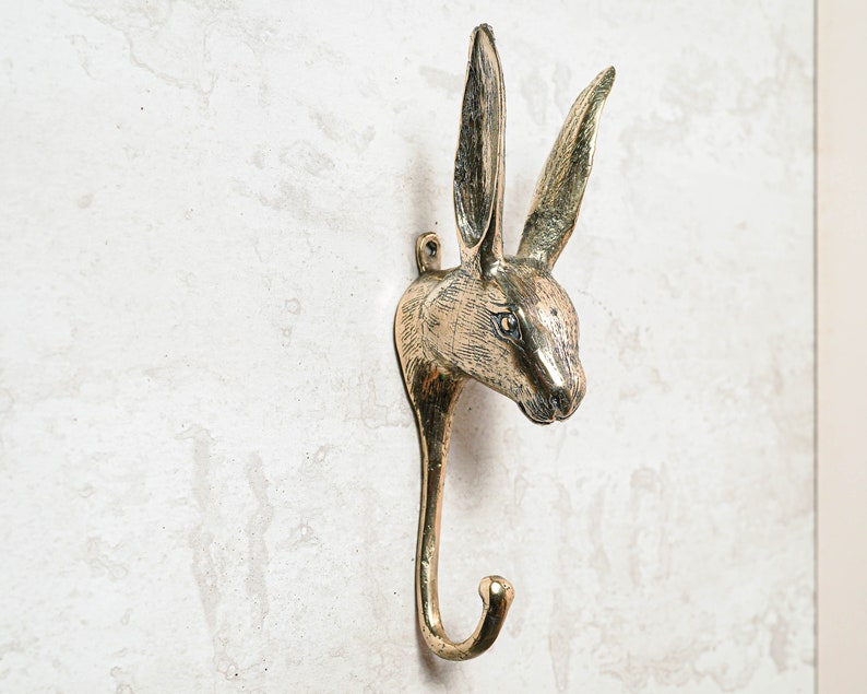 May include: A gold-toned metal rabbit head coat hook with a long, curved hook. The rabbit's ears are long and pointed, and its eyes are detailed.