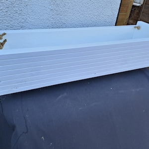 May include: A white wooden planter box with horizontal slats and rope handles. The rectangular container is painted white and has a nautical aesthetic. The planter is designed for outdoor use and is suitable for flowers or herbs.