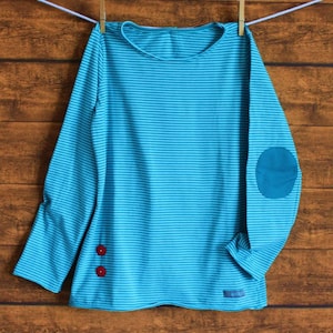 Könnte beinhalten: Ein türkisfarbenes, gestreiftes Langarmshirt mit einem Patch am rechten Ellenbogen und zwei roten Knöpfen auf der linken Seite.