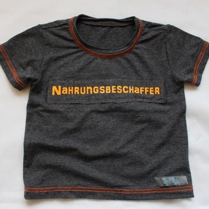 Könnte beinhalten: Ein dunkelgraues T-Shirt mit orangefarbenen Nähten an Ausschnitt und Ärmeln. Das Shirt hat einen gelben Aufnäher mit dem Text "Nahrrungsbeschaffer" auf der Vorderseite. Das Shirt hat ein kleines blaues Etikett mit dem Text "art-ich" unten rechts.