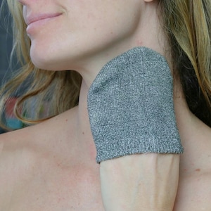 Puede incluir: Un guante exfoliante gris se sostiene contra el cuello de una persona. El guante está hecho de una tela texturizada, probablemente para el cuidado de la piel. El guante es de color gris liso.