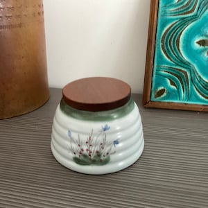Może przedstawiać: Biała ceramiczna miska z drewnianym wiekiem i kwiatowym wzorem. Miska ma zielono-biały pasiasty wzór i przedstawia niebieskie kwiaty i czerwone jagody.