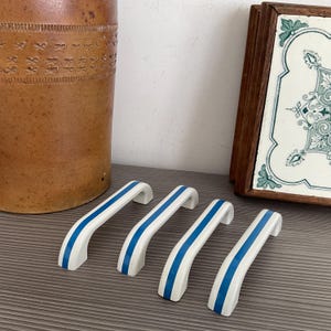 Puede incluir: Cuatro pomos de armario de cerámica blanca con rayas azules. Los pomos están dispuestos en fila sobre una superficie gris.