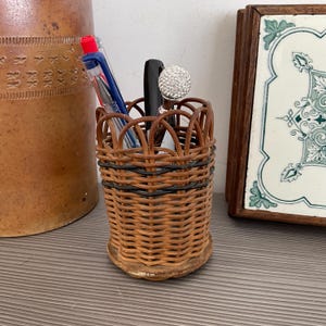May include: A small, woven wicker pen holder with a brown and black design. The holder contains pens and a decorative pen with a crystal-like top. A ceramic jar and a framed tile are in the background.