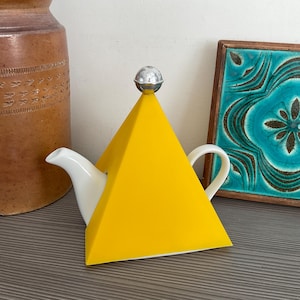 Può includere: Una teiera bianca con un coperchio giallo a forma di piramide e una sfera argentata sulla parte superiore. La teiera ha un beccuccio e un manico curvi. Lo sfondo include un vaso di ceramica marrone e una piastrella turchese incorniciata.