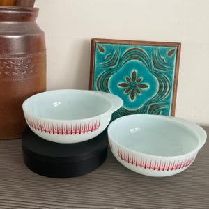 May include: Two white Pyrex bowls with red flame-like designs. The bowls have handles and are stacked on a black surface. A brown ceramic jar and a turquoise tile with a floral design are in the background.