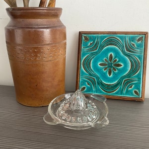 May include: A brown ceramic jar with a decorative pattern, a teal blue and brown ceramic tile with a floral design, and a glass lemon squeezer.