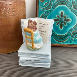 Puede incluir: Un sujetalibros de cerámica azul y blanco con un personaje de ratón sentado en una silla y leyendo un libro. El sujetalibros tiene el texto "Hunca Munca has got the cradle, and some of Lucinda's wishes." en el libro. El sujetalibros está sobre una superficie de madera.