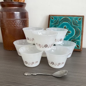 Puede incluir: Conjunto de seis tazas de cerámica blanca para pudín con un diseño floral marrón. Las tazas están apiladas en forma de pirámide sobre una superficie gris.