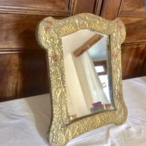 May include: An ornate gold-coloured mirror with a decorative floral design. The mirror is rectangular in shape and framed with a detailed, embossed pattern.