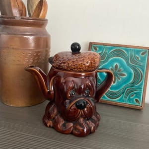 May include: A brown ceramic teapot shaped like a dog's head with a brown lid. The teapot has a black knob on the lid and a brown and black dog's face.