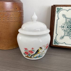 Op de afbeelding: Witte keramische pot met een deksel, versierd met een bloemenmotief en een gele vogel met zwarte accenten. De pot heeft een ronde vorm en een gladde, glanzende afwerking.