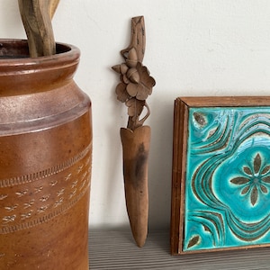 May include: A wooden letter opener with a carved acorn and leaf design. The opener is brown and has a pointed tip. It is sitting on a table next to a brown ceramic jar and a framed blue and green tile.