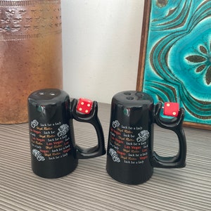 Könnte beinhalten: Zwei schwarze Salz- und Pfefferstreuer aus Keramik mit einem Würfeldesign am Griff. Die Streuer haben den Text "Las Vegas" und "Luck be a Lady" aufgedruckt.
