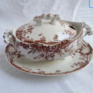 May include: A white porcelain gravy boat with a brown floral pattern and a lid. The gravy boat is on a matching plate.