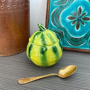 May include: A yellow and green ceramic squash-shaped jar with a lid, and a gold spoon. The jar has green vertical stripes and a small stem on top. The spoon is gold and has a decorative handle.