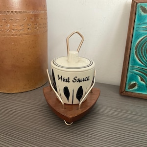 Może przedstawiać: Biały ceramiczny słoik z czarno-białym wzorem liści i napisem "Mint Sauce" na boku. Słoik ma metalową pokrywkę z uchwytem i stoi na drewnianej podstawce z trójkątnym dnem.