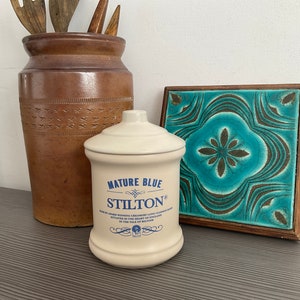 May include: A white ceramic jar with a blue label that reads "Mature Blue Stilton" and a decorative design. The jar is on a gray surface next to a brown vase and a blue and brown tile.