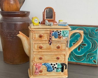 May include: A ceramic teapot shaped like a dresser with five drawers. The teapot is decorated with a mirror, a clock, and clothing items. The teapot is brown with gold accents.