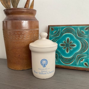 May include: A white ceramic jar with a blue and white label that reads "Mature Blue Stilton Cheese" and a blue ribbon with a crown. The jar is sitting on a wooden surface next to a brown ceramic vase and a blue and green tile.