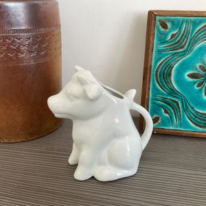 Peut inclure: Une crémière en céramique blanche en forme de vache, avec une anse et un bec verseur. Elle est posée sur une surface grise, avec un vase marron et une tuile turquoise encadrée en arrière-plan.