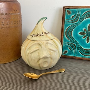 May include: A ceramic pickled onion jar with a face design, a green stem, and the words "Pickled Onion" in black script. A gold spoon rests in front of the jar. A brown ceramic jar and a framed turquoise tile are in the background.