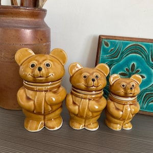 May include: A set of three graduated ceramic bear canisters with glossy honey-brown glaze. Each bear features a smiling face, rounded ears, and a sculpted scarf around the neck. The canisters are arranged in a row from largest to smallest.