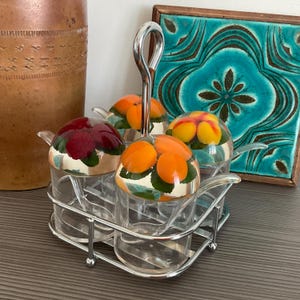 May include: A condiment server set featuring four clear glass jars with matching lids, each topped with a different fruit motif including red berries, orange citrus, and yellow peaches, all held in a chrome metal caddy with a central handle.