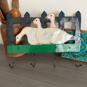 May include: A decorative wall-mounted key holder featuring three white geese painted on a green and black wooden fence. The key holder has three metal hooks for hanging keys or other small items. The geese have orange beaks.