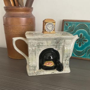 Puede incluir: Una tetera de cerámica blanca con forma de chimenea con una chimenea negra y un reloj dorado en la parte superior. La tetera tiene un diseño de mármol y una figura de gato negro sentada frente a la chimenea.