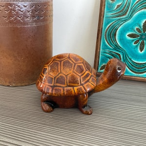 May include: A ceramic turtle ornament with a brown and orange shell design. The turtle has a long neck and small legs. It is placed on a grey surface. The figurine is a decorative item, likely for a home.