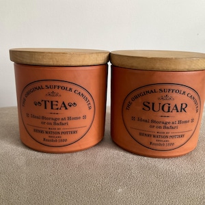 May include: Two orange ceramic canisters with wooden lids. One reads "TEA" and the other "SUGAR" in a circular design. The text "The Original Suffolk Canister" is also visible.
