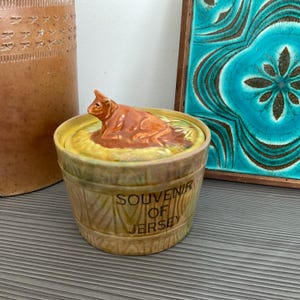 May include: A vintage ceramic souvenir jar from Jersey, featuring a brown cow figurine on the lid. The jar has a barrel-like design with the words "Souvenir of Jersey" in black lettering. The jar is tan and yellow.