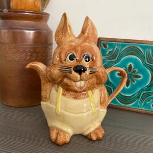 May include: A brown and tan ceramic teapot shaped like a rabbit wearing overalls. The rabbit has a wide smile and large eyes. The teapot has a spout and a handle.