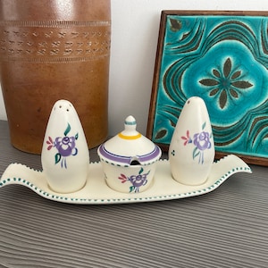 May include: A white ceramic salt and pepper shaker set with a floral design in shades of purple, pink, and green. The shakers are on a white ceramic tray with a green trim. There is a small white ceramic dish with a purple and yellow trim in the center of the tray.