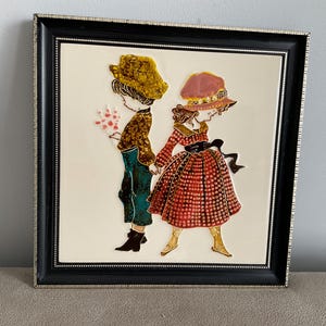 Könnte beinhalten: Gerahmte Keramikfliesenkunst mit zwei Figuren, die sich an den Händen halten. Eine Figur trägt einen gelben Hut und eine braune Jacke und hält Blumen. Die andere Figur trägt einen rosa Hut und ein rotes Kleid. Das Kunstwerk ist schwarz gerahmt mit silbernem Rand.