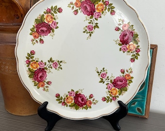 May include: A white ceramic serving platter with a scalloped edge and a gold rim. The platter is decorated with a floral pattern of pink, yellow, and green roses.
