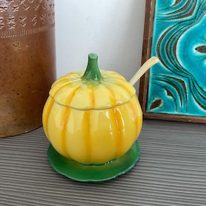 May include: A yellow ceramic pumpkin-shaped sugar bowl with a green stem and lid, on a green saucer. A small spoon is inside. The bowl has vertical yellow stripes. Decorative kitchenware, with measurements in cm.
