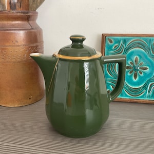 May include: A dark green ceramic teapot with a gold trim around the lid and spout. The teapot has a rounded body and a slightly flared spout.